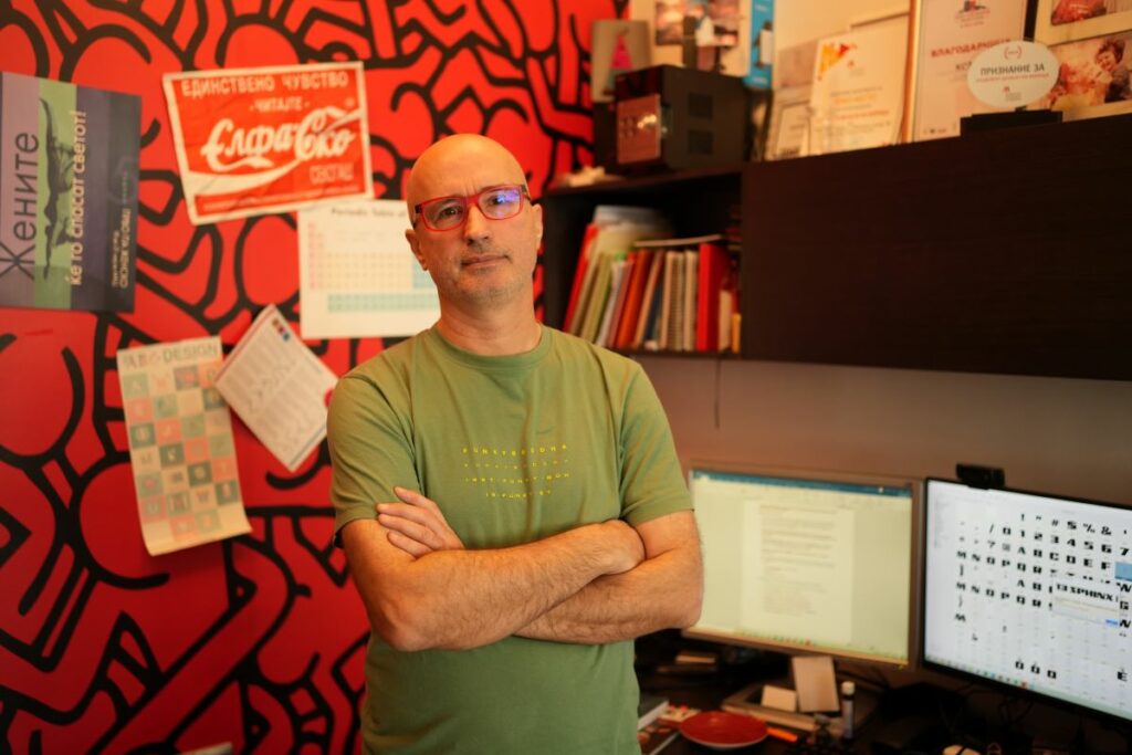 Небојша Гелевски – Бане стои со скрстени раце во своето работно студио, пред компјутери со прикажани кирилични фонтови, со црвена ѕидна позадина исполнета со графички постери и признанија.